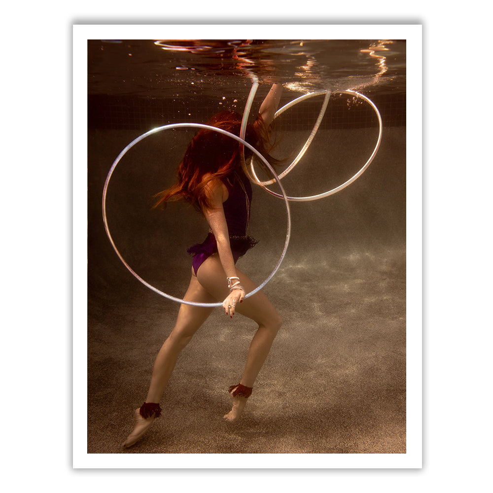 Dressed in a purple ensemble, a performer dances with two glowing hoops underwater, where the light casts a reddish hue and the pool's bottom remains visible. The scene evokes the fluid motion and elegance of fine art paper and feels as if it were custom made for appreciation. Experience this captivating moment with Francesca Owen’s Show Time 4.