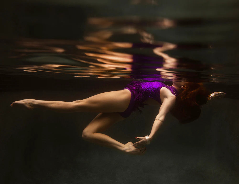 Wearing the Show Time Part 2, #2 by Francesca Owen, a person floats underwater with graceful movements, their reflection creating a serene, artistic effect. The calm water serves as a tranquil backdrop reminiscent of fine art paper, capturing the moment as if crafted with archival pigments.