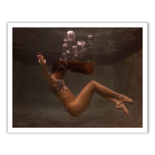 Wearing the exquisitely designed Show Time Part 3, #4 bikini by Francesca Owen, a person with long hair floats gracefully underwater. Bubbles ascend around them, capturing their mid-motion elegance in a dimly lit, serene swimming pool environment reminiscent of a fine art paper's depiction of tranquil beauty.