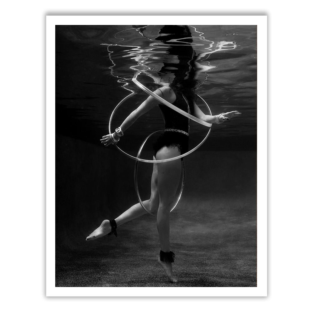 Amidst a surreal underwater scene, Show Time 6 by Francesca Owen captures a person elegantly hula hooping. Encircled by hoops and wearing a black swimsuit, their reflections dance on the water's surface. This captivating black-and-white image is printed using archival pigments, hand-signed by the artist, and comes with a certificate of authenticity.