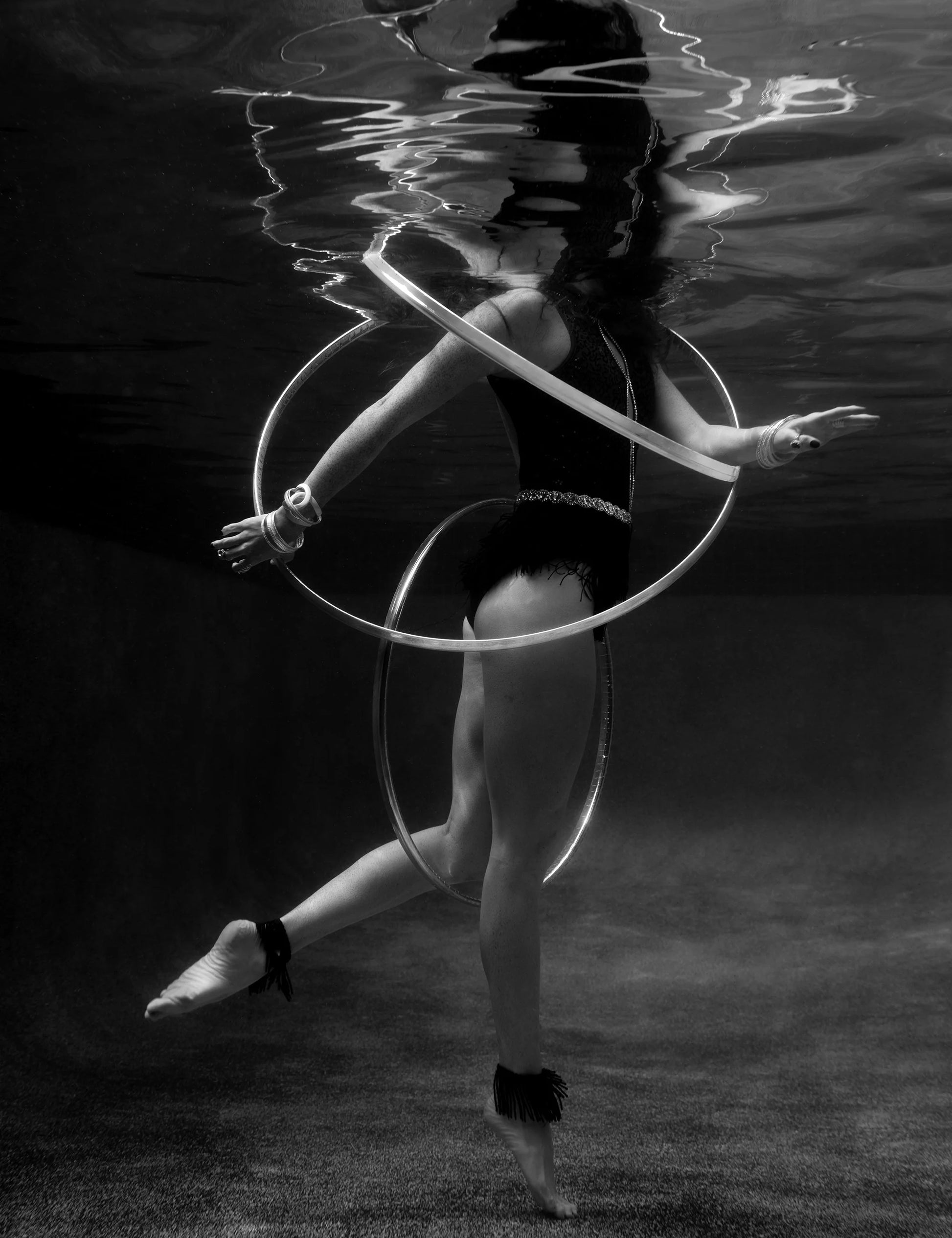The black and white underwater photograph, titled "Show Time 6," showcases a person in a swimsuit elegantly hoop dancing. Their hair floats upward as they skillfully spin multiple hoops around their arms and waist. Captured using archival pigments, this artwork by Francesca Owen is hand-signed and comes with a certificate of authenticity.