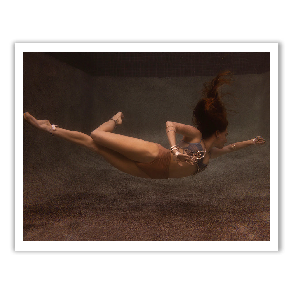 A woman swims gracefully underwater, her long hair flowing behind her. Captured by Francesca Owen, she showcases "Show Time Part 3, #6" in a bikini and jewelry set against a soft, sandy background. The lighting creates an ethereal atmosphere, ideal for reproduction with archival pigments on fine art paper.
