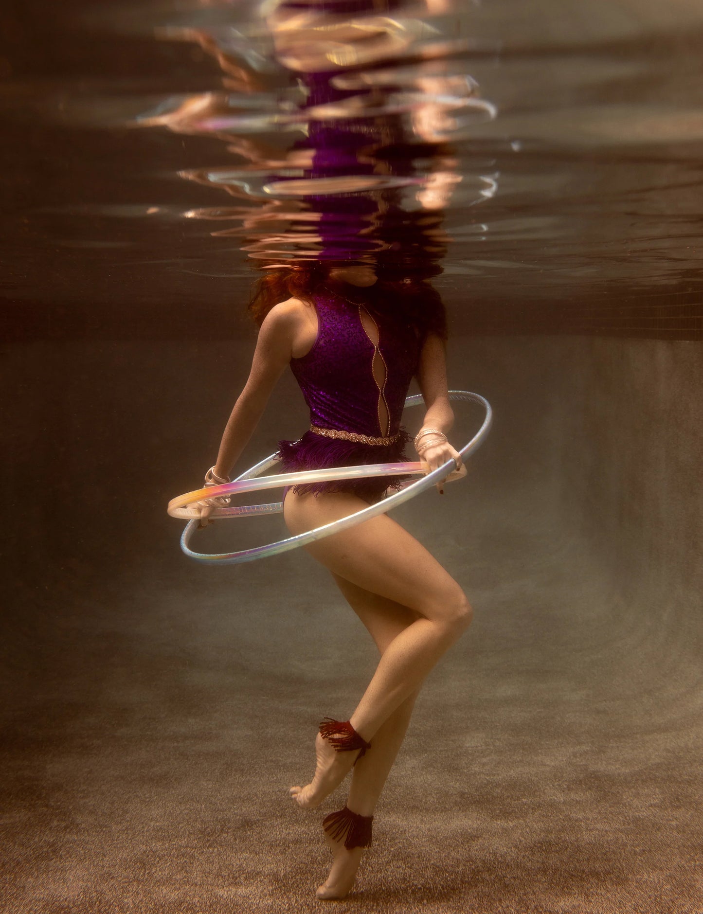A person is performing underwater hula-hooping with the Show Time 7, a glowing hoop by Francesca Owen, dressed in a vibrant purple outfit with long hair flowing. The water distorts their reflection above, giving the scene an ethereal quality akin to fine art paper prints created with archival pigments.