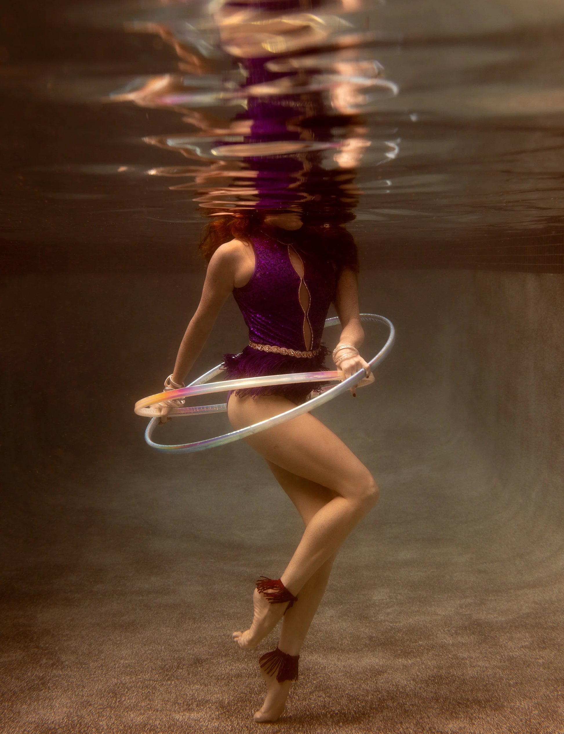 A person is performing underwater hula-hooping with the Show Time 7, a glowing hoop by Francesca Owen, dressed in a vibrant purple outfit with long hair flowing. The water distorts their reflection above, giving the scene an ethereal quality akin to fine art paper prints created with archival pigments.
