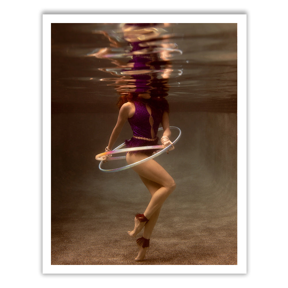 The image "Show Time 7" by Francesca Owen presents a dream-like underwater scene where a person in a purple outfit hula-hoops. The reflective water beautifully enhances the setting, showcasing the sandy pool floor. Printed on fine art paper with archival pigments, this artwork comes with a certificate of authenticity.