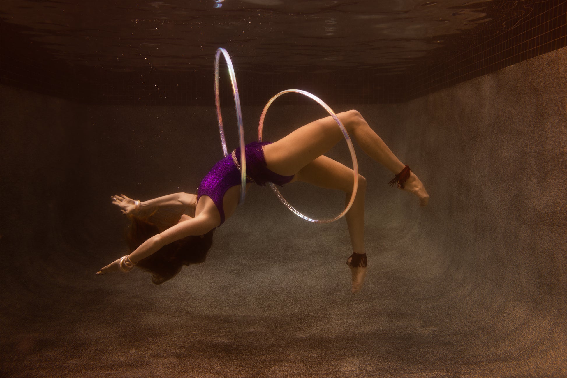 A person in a purple outfit performs an underwater dance routine in a swimming pool, gracefully arching backward while manipulating several glowing hula hoops around their body. The scene, perfect for fine art prints, is captured in "Show Time 2" by Francesca Owen and is set against the textured, dark brown tiled interior of the pool.