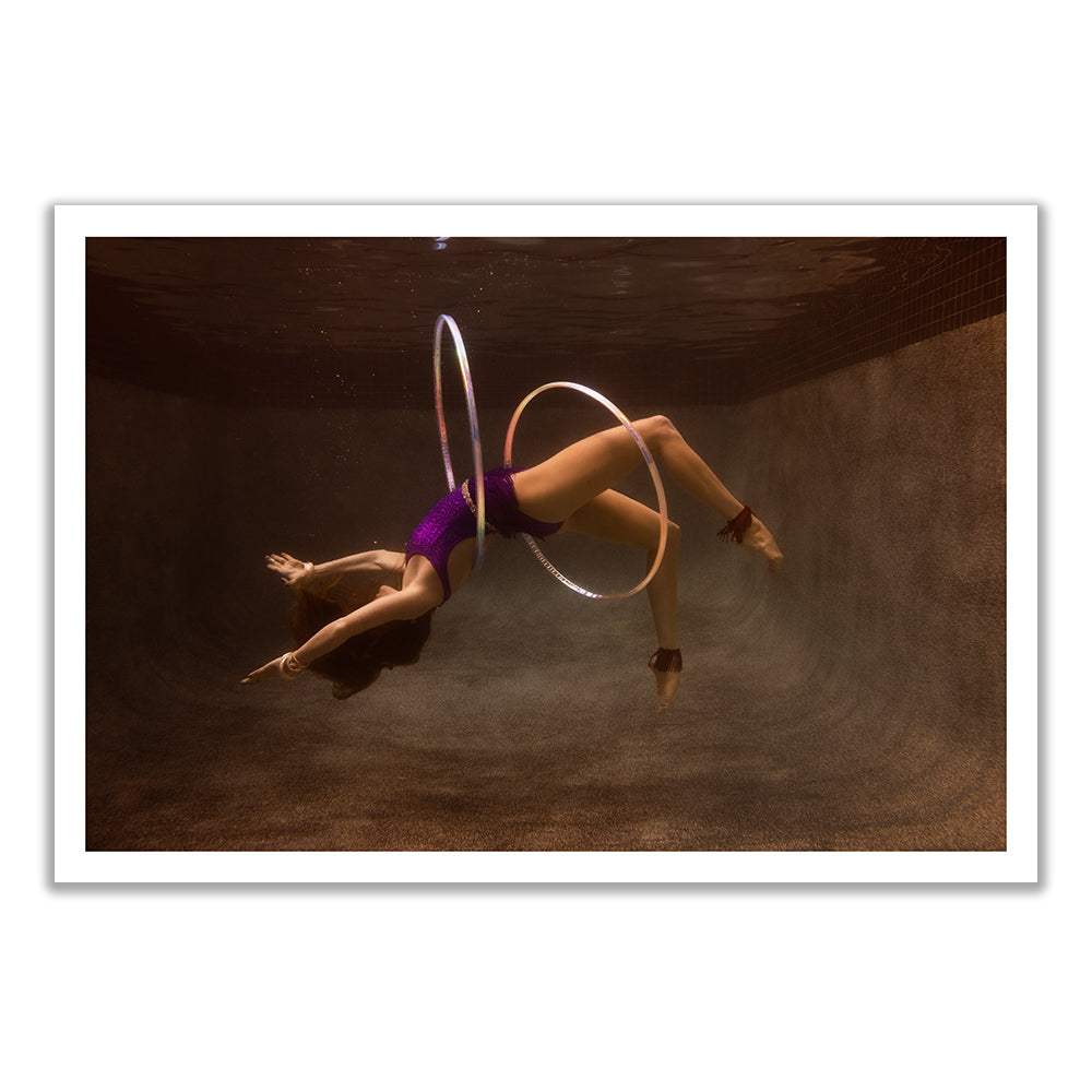 A person underwater, donned in a purple outfit and striking an acrobatic pose as they expertly balance two glowing hula hoops—one around their waist and another around their ankle. The textured walls of the swimming pool serve as the backdrop, beautifully captured with archival pigments for fine art prints in custom sizes. This captivating scene is from "Show Time 2" by Francesca Owen.