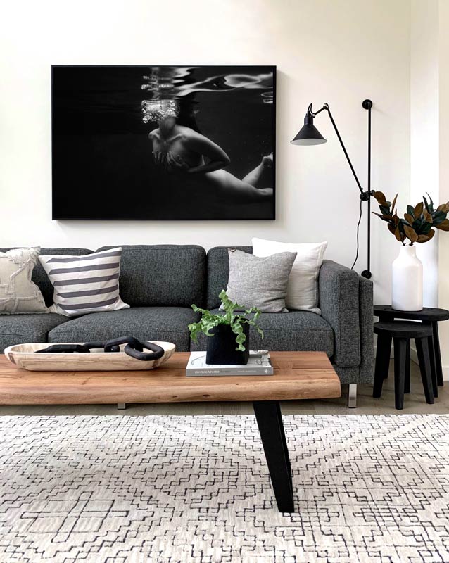 A modern living room featuring a gray sofa with assorted cushions, a wooden coffee table, and an area rug with geometric patterns. Hanging above the sofa is "Hold On," a large black-and-white artwork of an underwater scene by Francesca Owen, hand signed and printed on fine art paper. The room also includes a black floor lamp and a side table adorned with a vase.
