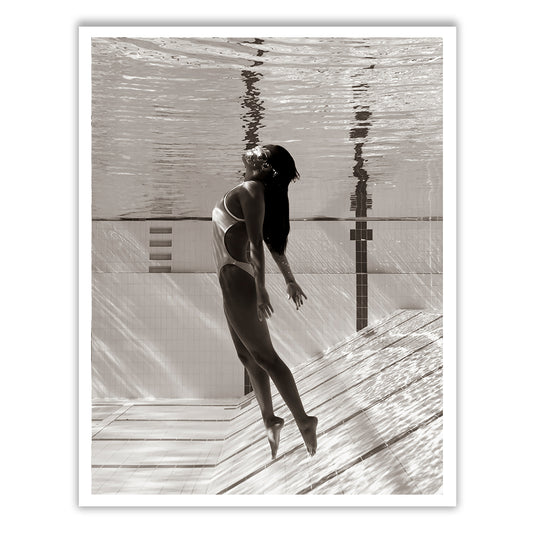 Flying Solo 05 by Francesca Owen captures a black-and-white underwater photograph of a woman in a one-piece swimsuit. She is submerged in a swimming pool, facing upward and appearing to float gracefully near the pool's edge where light patterns are visible on the water surface and pool tiles. Printed on fine art paper with archival pigments, this piece includes a certificate of authenticity.