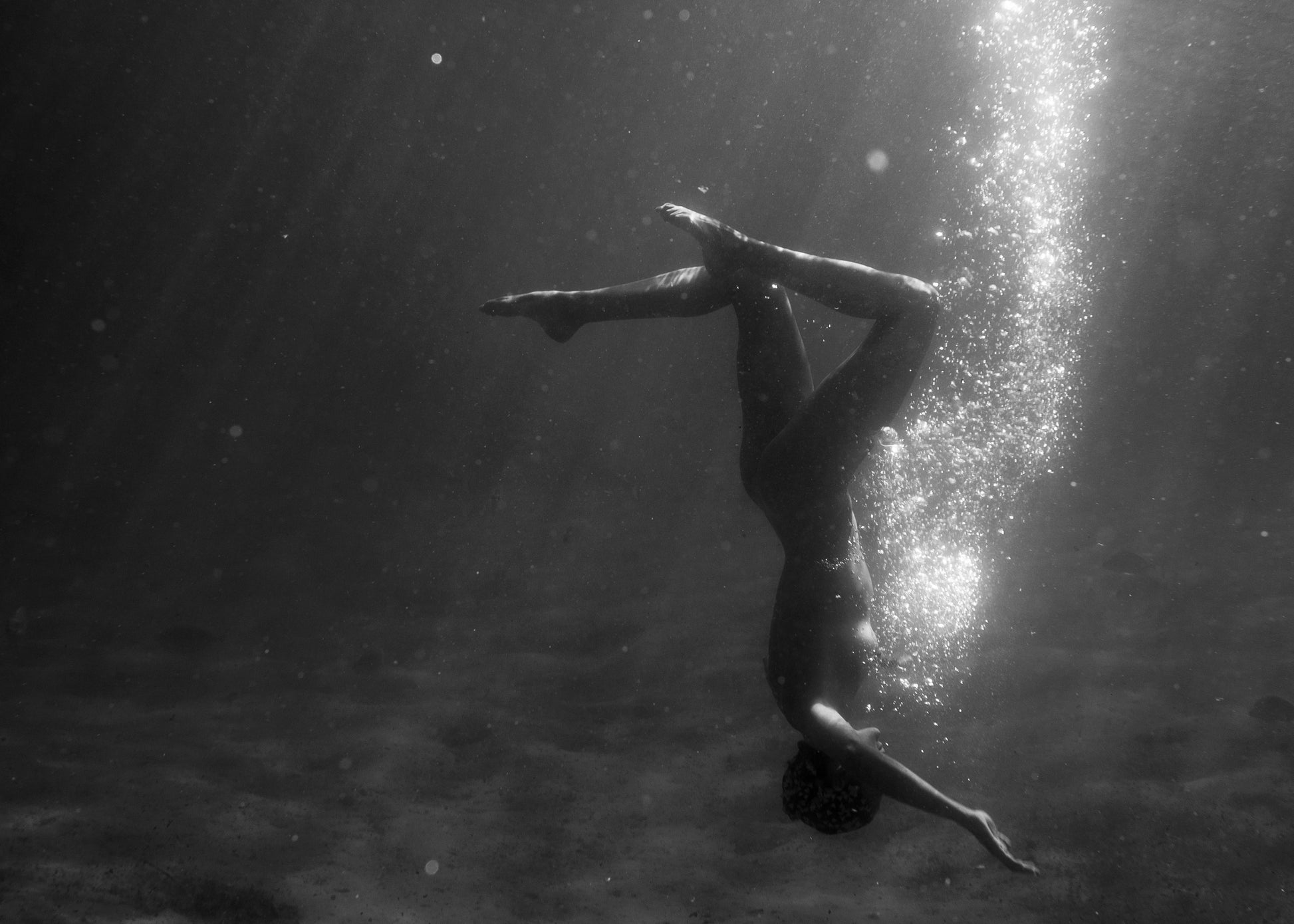 Still Waters #9 by Francesca Owen features a black-and-white image of a person underwater performing an upside-down pose with legs crossed and arms extended. Bubbles rise around them, while light beams penetrate the water, creating an ethereal atmosphere. This stunning piece is printed on fine art paper using archival pigments for lasting quality.