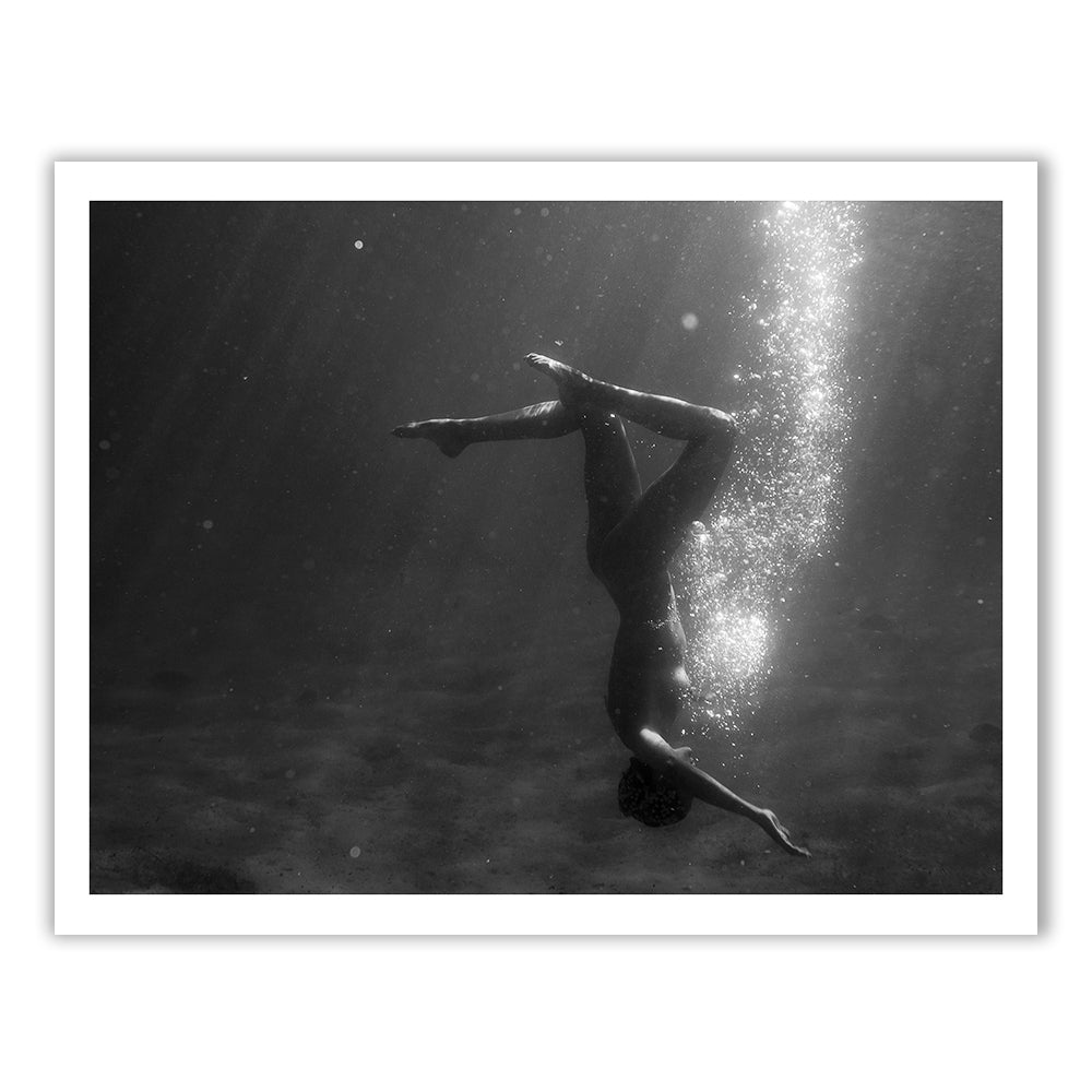 Still Waters #9 from Francesca Owen captures a black and white photo of a person underwater, gracefully upside down with legs bent and arms extended. Bubbles rise to the surface while light rays from above penetrate the water, creating an ethereal scene. Printed on fine art paper with archival pigments, it includes a certificate of authenticity.