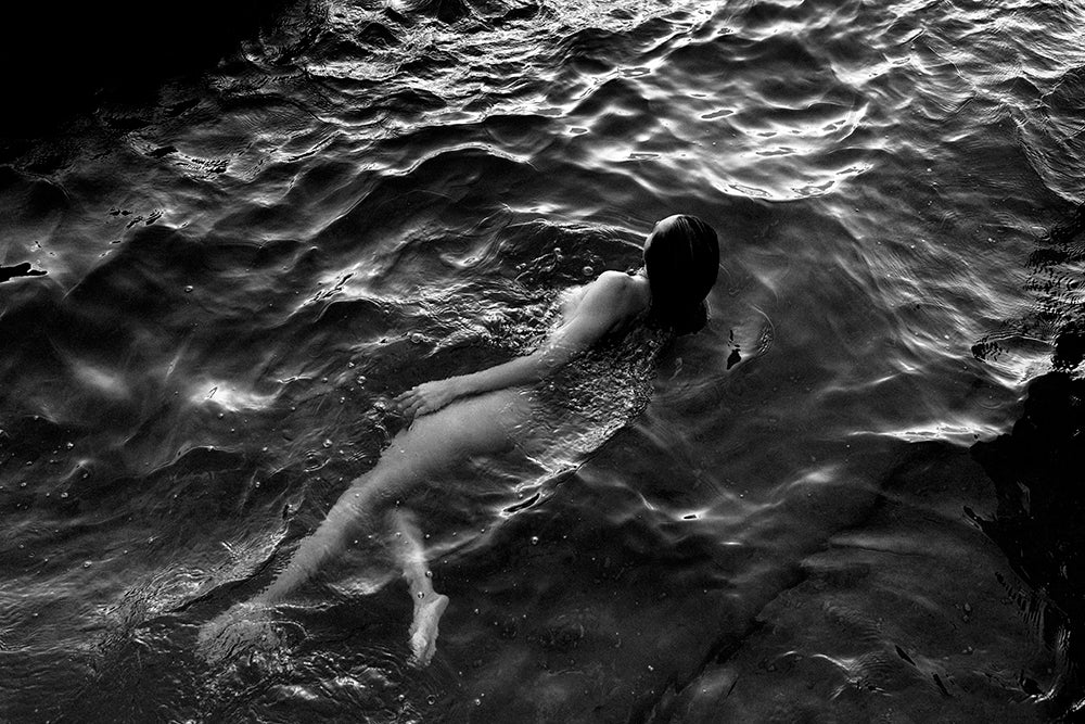 A black and white photo titled "Sunrise Swim #1" by Francesca Owen captures a person swimming in water. The swimmer's back is visible as they float and move through the rippling surface, surrounded by natural light and shadow reflections. Printed on fine art paper, each piece comes with a certificate of authenticity, with worldwide shipping available.