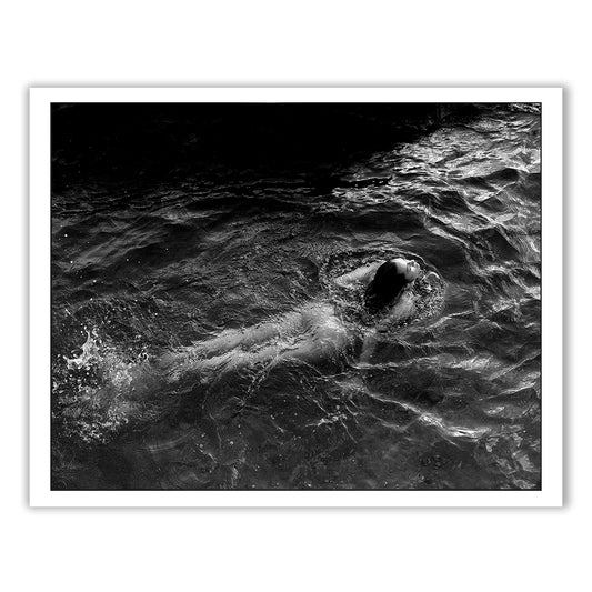 Product description for "Sunrise Swim #2" by Francesca Owen:
This black and white photograph, titled "Sunrise Swim #2" by Francesca Owen, captures a person swimming on their back in rippling water. The light casts intricate shadows that emphasize the texture of the water and highlight the swimmer's relaxed posture. Printed with archival pigments on fine art paper, this image encapsulates a serene moment of calm.