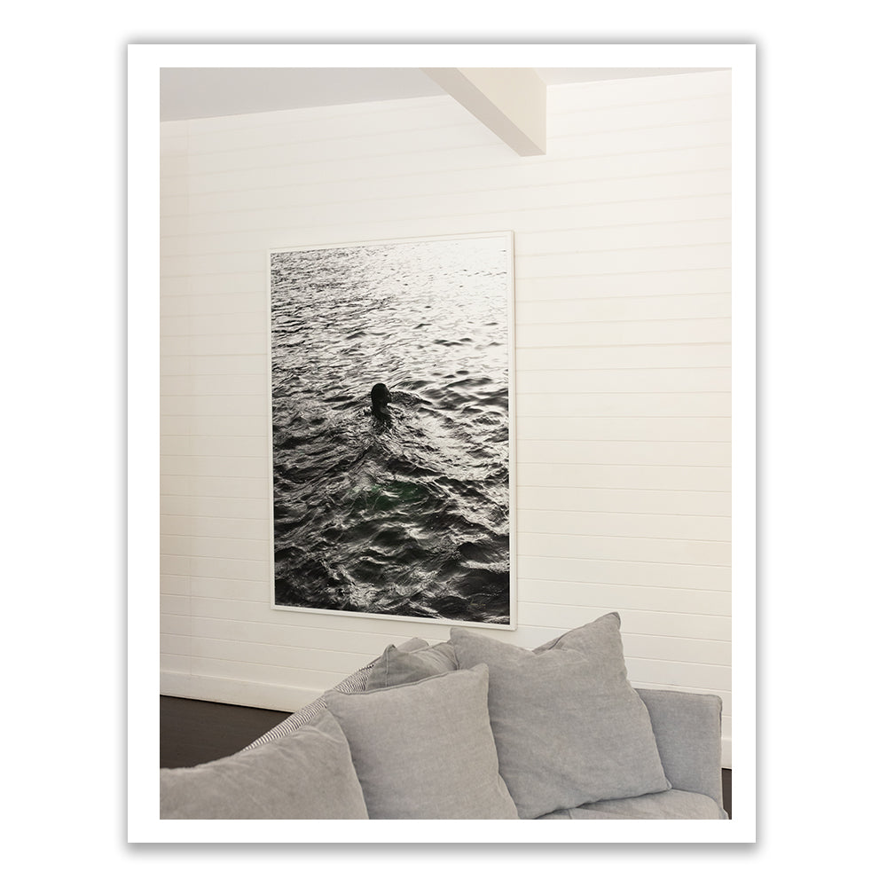 A framed photograph entitled "Sunrise Swim #3" by Francesca Owen adorns the white wall, depicting a person swimming in water. Positioned in front of this artwork is a sofa with light gray cushions, creating a serene and custom-made ambiance.