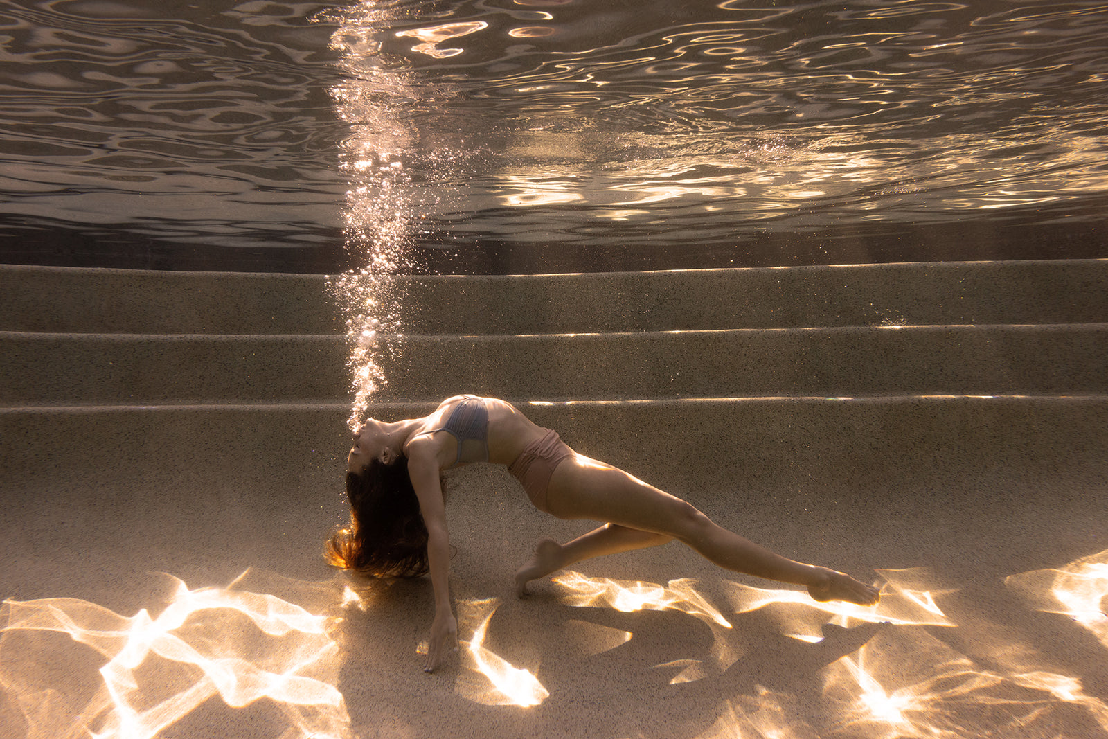 A woman in a swimsuit arches gracefully underwater on pool steps, exhaling bubbles as sunlight patterns shimmer below. "Sunset #2," a fine art print by Francesca Owen, includes a certificate of authenticity.