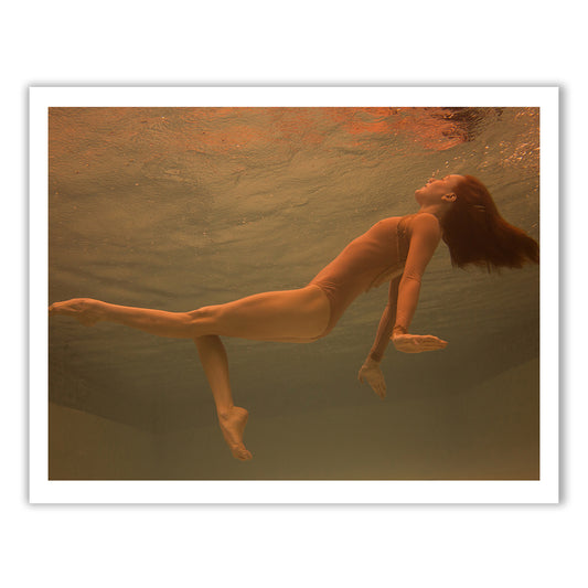 The Francesca Owen "Surfacing" features a woman in a one-piece swimsuit underwater, floating gracefully with her arms extended and legs posed. The scene, reminiscent of fine art prints, is bathed in warm, reddish light, creating a serene and ethereal ambiance as her hair flows freely in the water.