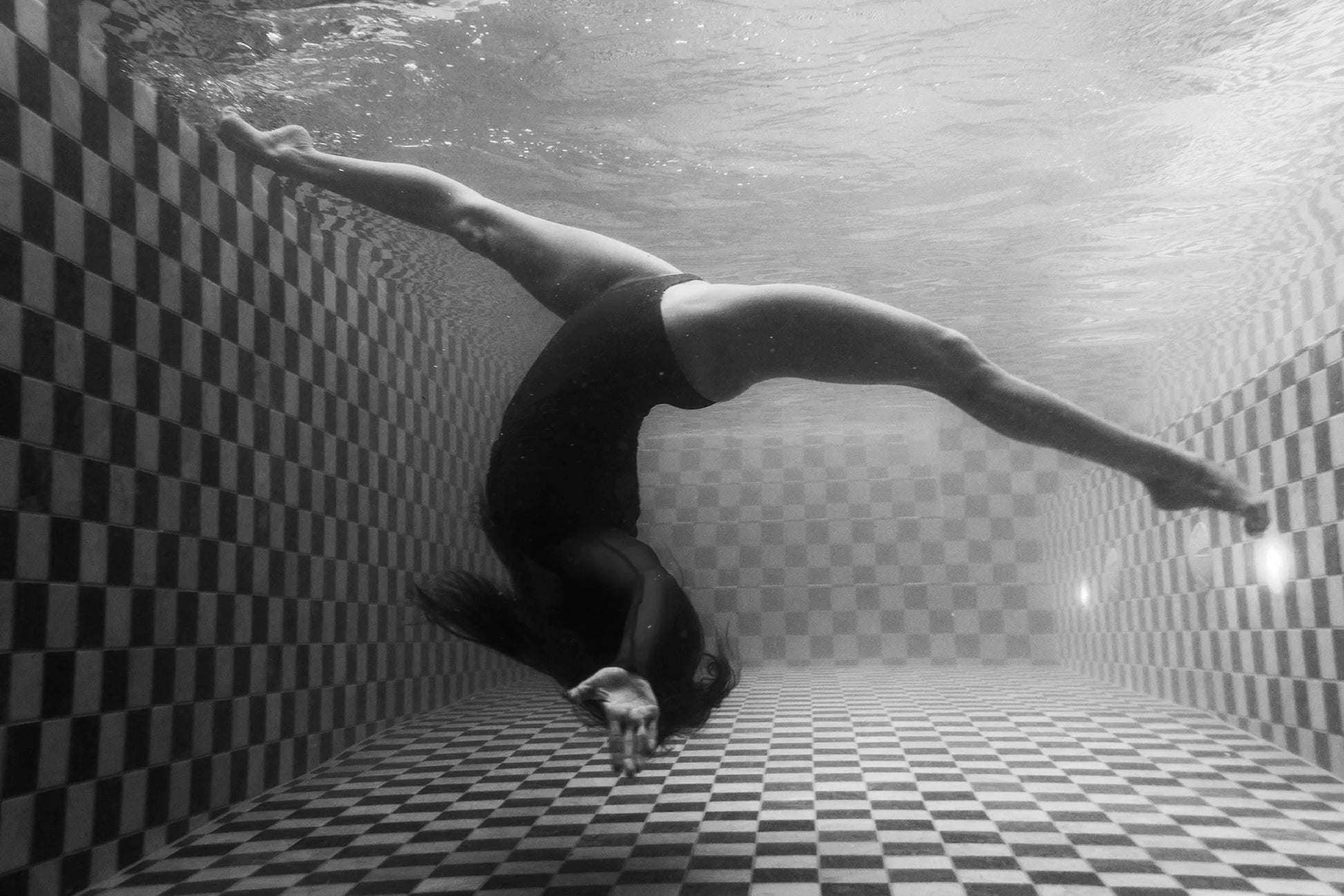 Wild at Heart no.4 B&W by Francesca Owen features a black-and-white image of a person underwater in a pool, performing an acrobatic pose. The individual is arched backward with legs spread wide, touching the tiled pool floor with their hands. The pool showcases a checkered pattern on the floor and walls. This striking piece is available in custom sizes printed on fine art paper.