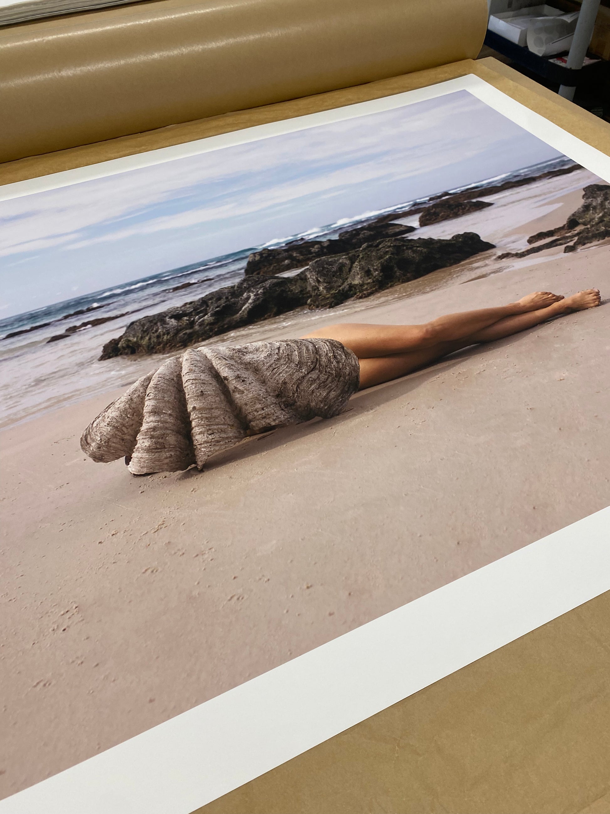 A photo titled "The Beauty Within" by Francesca Owen features a person lying on a sandy beach with their lower body visible and their upper body covered by a large clamshell. Waves and rocks frame the background under a clear sky. This captivating image, printed with archival pigments on fine art paper, is available for worldwide shipping.