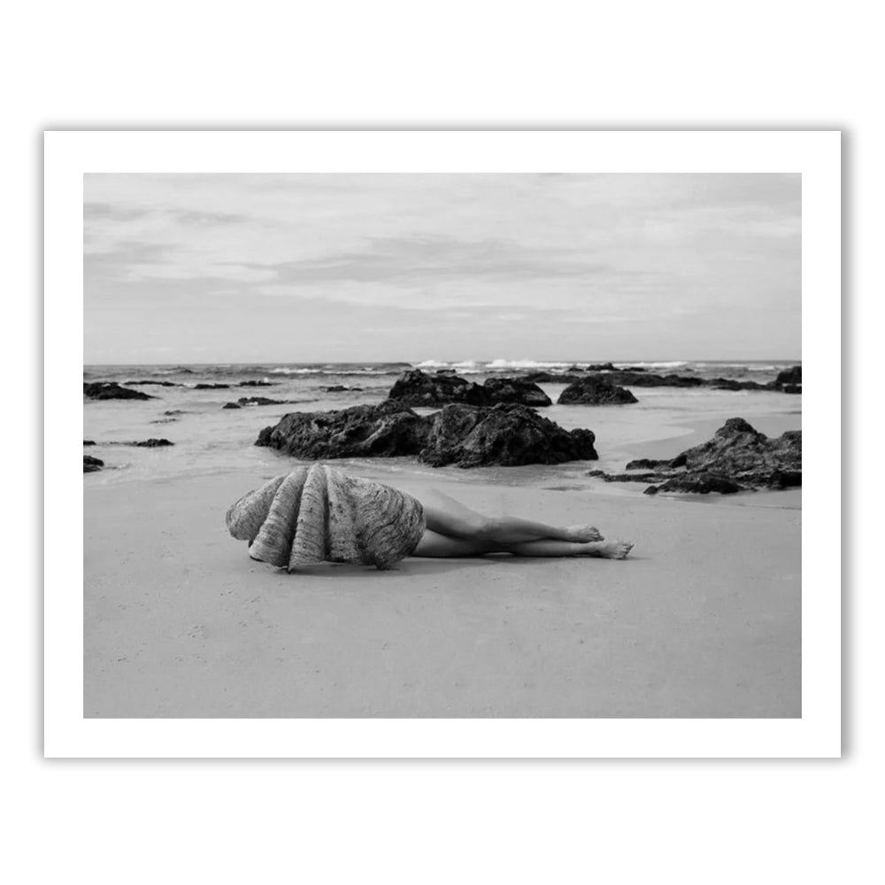 The Beauty Within B&W" by Francesca Owen captures a striking black-and-white image of a person lying on a beach with their upper body enveloped in a large seashell, set against rocky formations and a tranquil sea beneath a cloudy sky. This fine art photograph is now available with worldwide shipping.