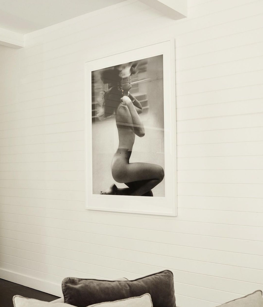 An artful black-and-white photograph from the "UNVEILED" collection by Francesca Owen, printed on fine art paper with archival pigments, graces a white shiplap wall. The image captures a partially clothed person in a seated partial profile. Below this framed Limited Edition print, a sofa adorned with grey and white cushions is partially visible.