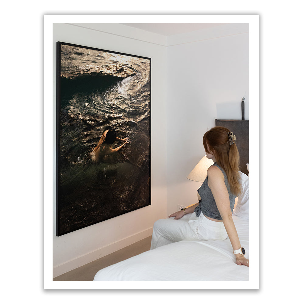 A person sitting on a bed in a modern, minimalist room gazes at "Sunrise Swim #9," a large framed photograph by Francesca Owen depicting someone swimming in dark, textured water. Mounted on the white wall and printed on fine art paper, this custom-made artwork is hand signed. A lamp on the bedside table softly illuminates the scene.