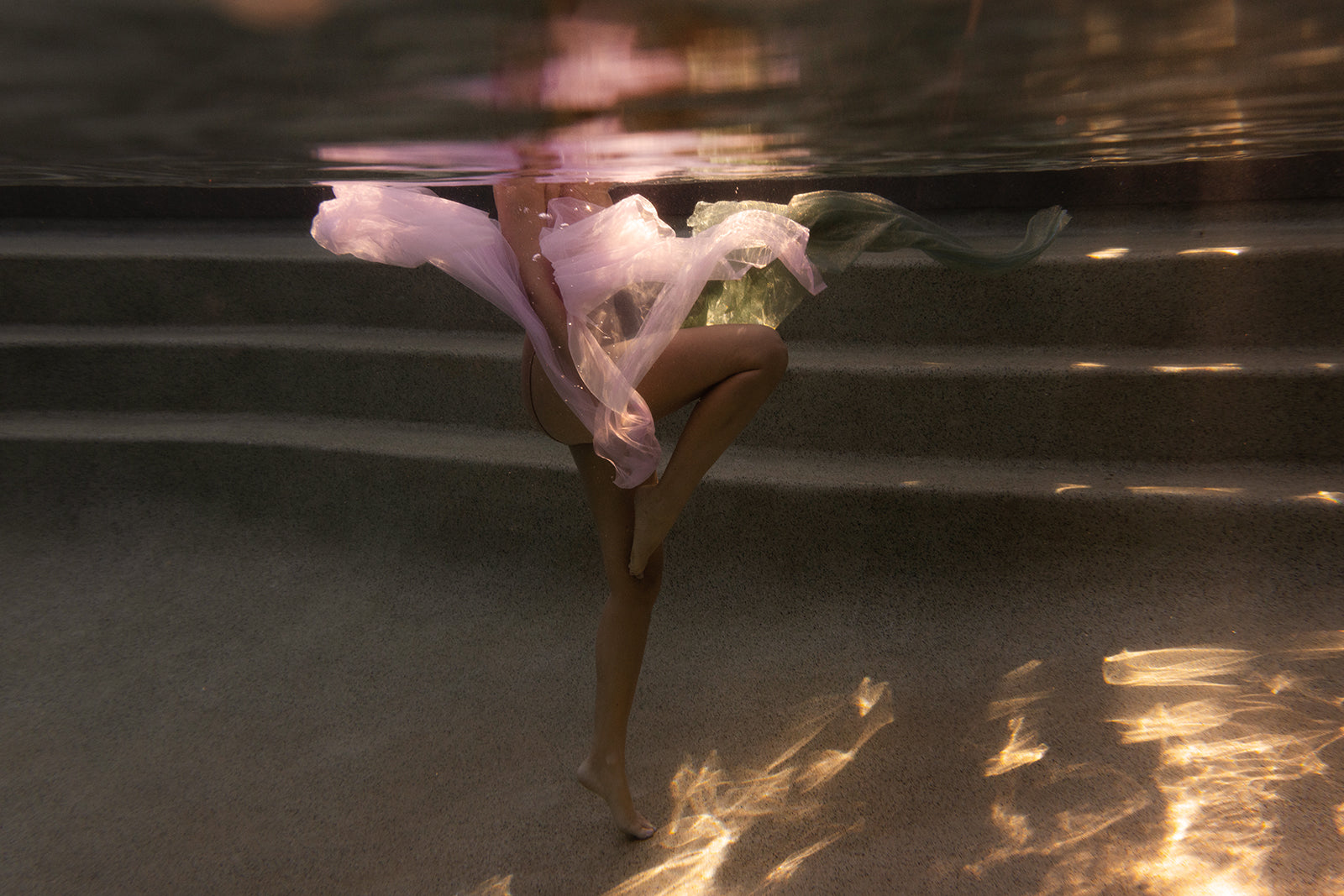Sunset #3 by Francesca Owen depicts a person underwater, wrapped in flowing sheer fabric, balancing on one leg as soft light and shadows play across the pool steps and their lower body in this evocative fine art print.
