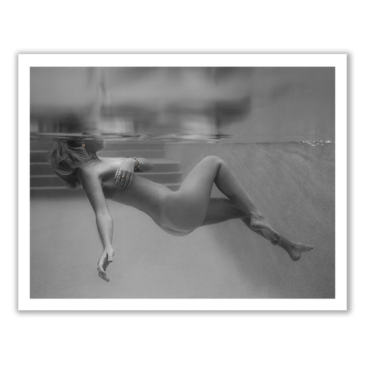 A person is floating underwater in a pool, partially submerged with their face obscured by the water's surface. The calm environment, highlighted by steps leading out of the pool in the background, captures a serene moment. This black and white image, titled "Unveiled 05" from Francesca Owen, exudes elegance and is perfect for fine art prints and custom framing.