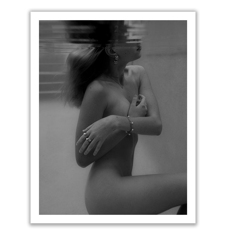 The "Unveiled 01" by Francesca Owen features a black and white photo of a nude individual partially submerged in water. The subject covers their chest with one arm while the other arm rests on their side, creating a striking division at the waterline that results in a reflective effect on their face. This piece is printed on fine art paper, hand signed by the artist, and includes a certificate of authenticity.