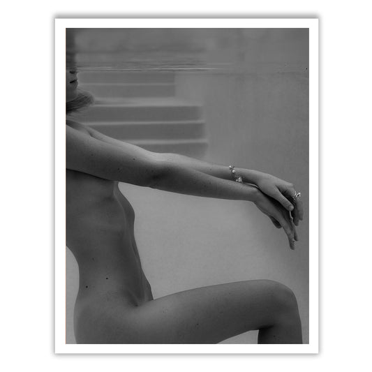 The photograph titled "Unveiled 03" by Francesca Owen features a person submerged in water up to their neck, with arms extended and hands clasped. Shadows play across their form, creating a serene and contemplative atmosphere. Rendered with archival pigments, this fine art print captures blurred steps underwater in the background.