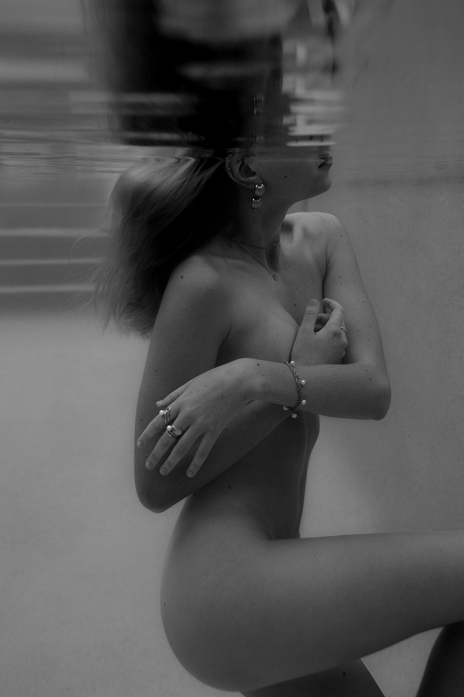 Black and white photo titled "Unveiled 01" by Francesca Owen depicts a person underwater with shoulder-length hair. They are holding their arms across their chest, partially covering themselves, and wearing several rings and bracelets. The person's face is partially out of the water. Printed on fine art paper, hand-signed with a certificate of authenticity included.