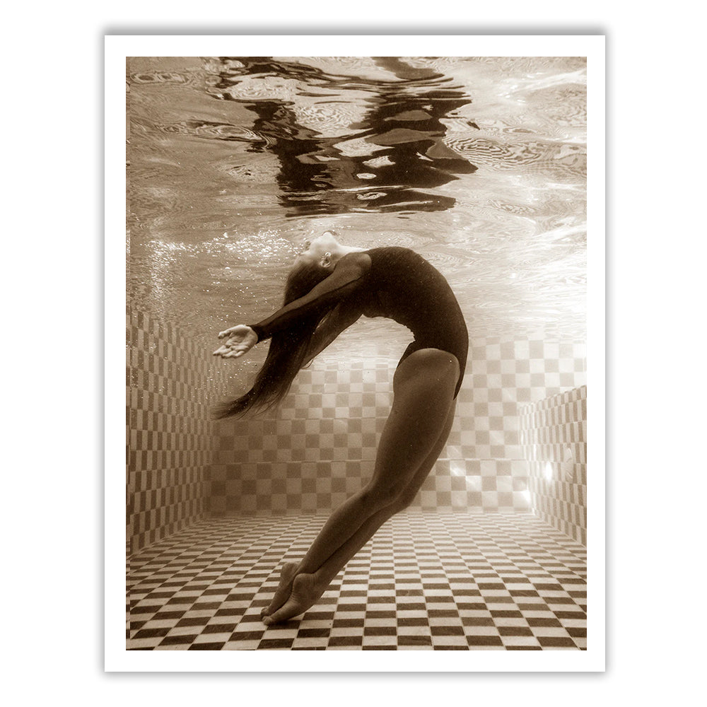 Wild at Heart no.1 by Francesca Owen features a sepia-toned image of a person underwater, arching backward gracefully with arms outstretched and long hair flowing. Standing on tiptoes in a checkered pool, they create a serene and elegant scene, captured on fine art paper using archival pigments and hand signed by the artist.