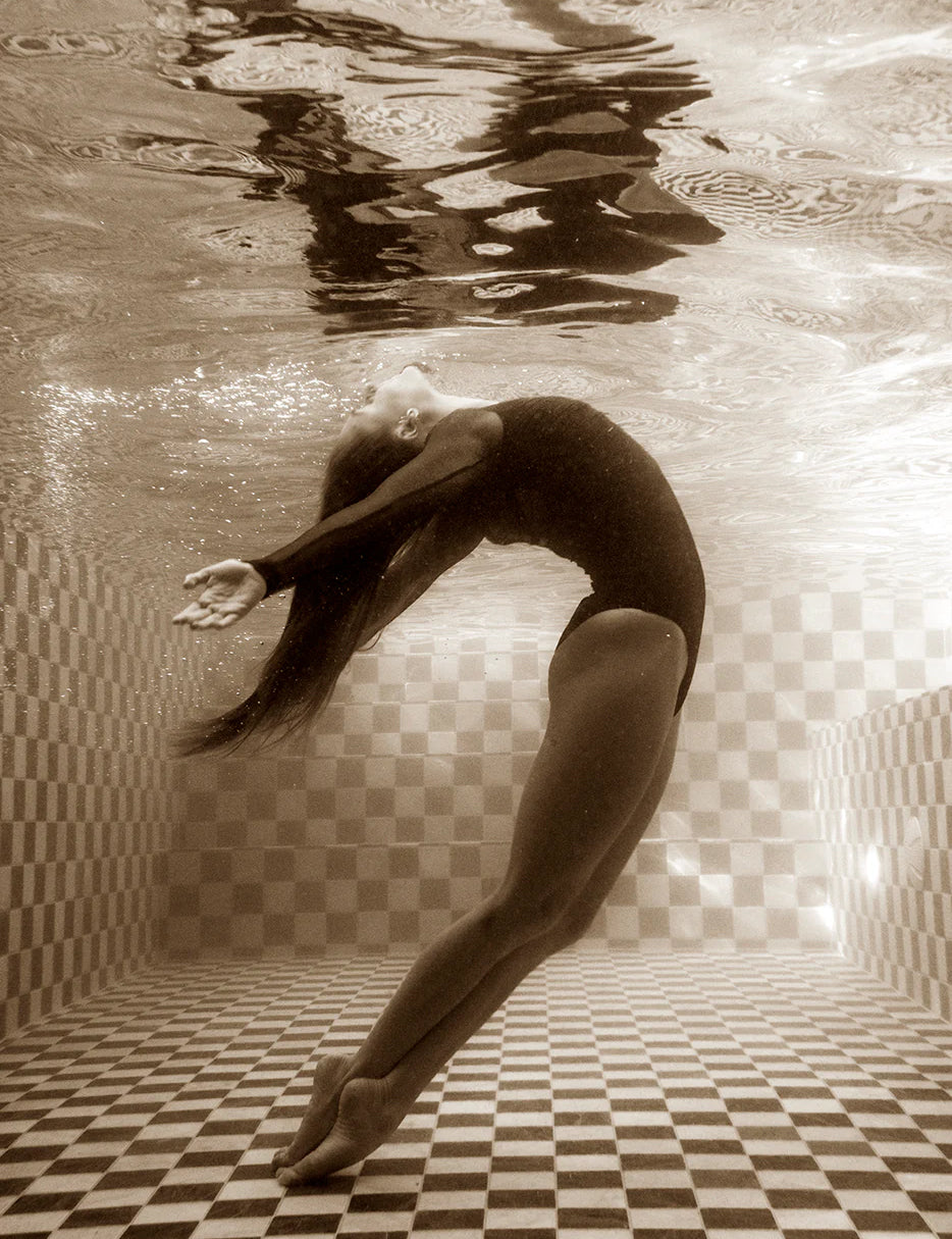 A person in a black swimsuit gracefully arches backward underwater in a checkered swimming pool, with long hair flowing freely. The sepia-toned image, titled "Wild at Heart no.1" by Francesca Owen, is printed on fine art paper and captures intricate reflections on the water's surface above. It is hand signed by the artist using archival pigments.