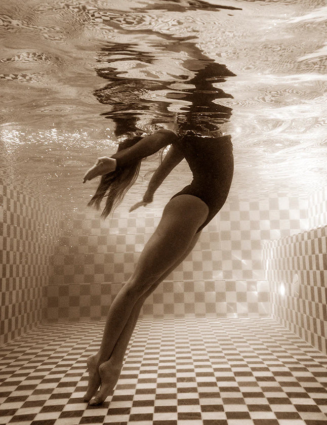 In "Wild at Heart no.2" by Francesca Owen, a sepia-toned underwater photograph showcases a person in a one-piece swimsuit poised gracefully with one arm extended upward and one leg backward near the water's surface. This fine art print, made with archival pigments, captures a serene moment in a tiled swimming pool adorned with a checked pattern.