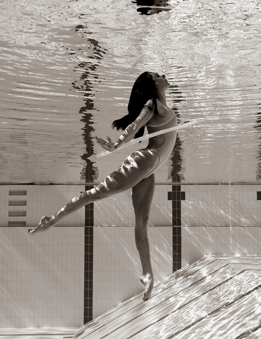In "Flying Solo 02" by Francesca Owen, a person in a swimsuit gracefully poses underwater in a pool, balancing on one leg with the other extended behind them. Their arms are outstretched, and their hair flows upwards. The water's surface ripples above, creating shimmering dappled light patterns. Printed on fine art paper using archival pigments, this stunning image comes with a certificate of authenticity.