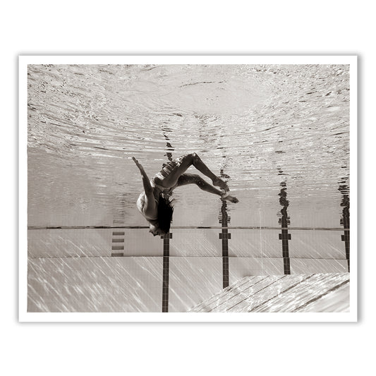 Flying Solo 07 by Francesca Owen is a black and white underwater photograph depicting a person floating upside down in a swimming pool. The individual's hair gracefully flows upward as they are submerged, with pool tiles and water reflections visible. This captivating image is captured in archival pigments on fine art paper, ensuring lasting beauty.
