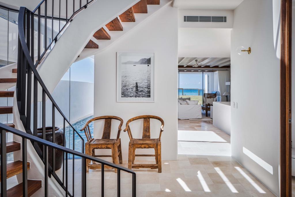 A modern interior features a staircase with wooden steps and a black railing on the left. Two Francesca Owen Capri Tippy Toes wooden chairs are positioned against a white wall adorned with a black-and-white photo printed with archival pigments. Bright natural light streams into the room, which leads to a living area with an ocean view.