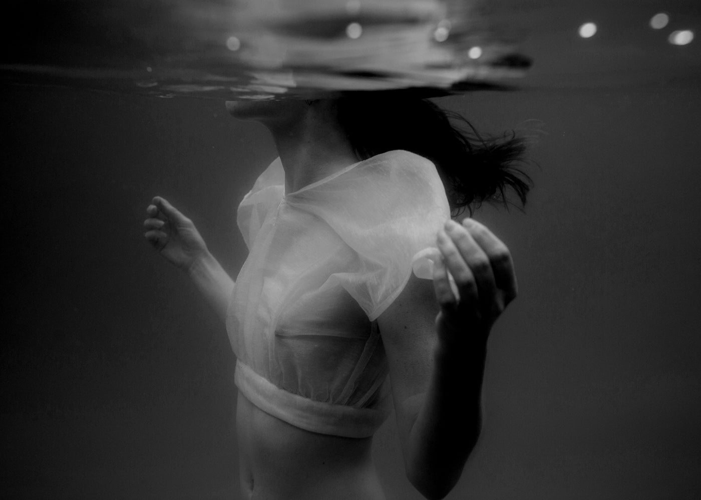Accepting" by Francesca Owen is a black-and-white underwater photograph capturing a person with long hair partially submerged, wearing a sheer, light-colored garment. They appear to be gazing upwards, surrounded by bubbles that contribute to the dreamlike and ethereal ambiance. This evocative artwork is printed on fine art paper using archival pigments for enduring quality.