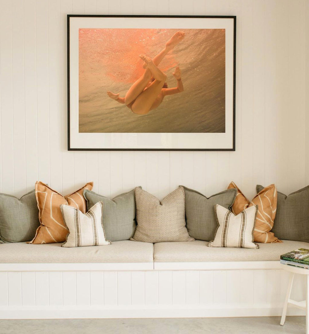 A cozy seating area features a built-in white bench adorned with various gray and brown pillows. Above the bench, a framed, hand-signed photograph titled "Another World" by Francesca Owen, depicting a person swimming underwater bathed in warm light, hangs on a beadboard wall. A small white table is nearby.