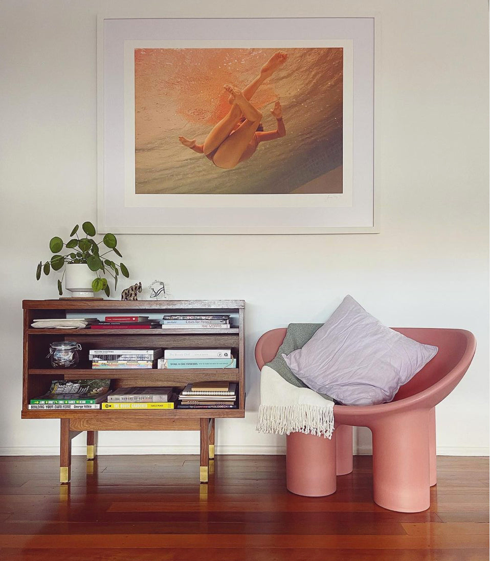 A cozy interior features a modern pink chair, draped with a blanket and pillow, positioned next to a wooden bookshelf adorned with an array of books and decor. Above the shelf hangs "Another World," a hand-signed photograph from Francesca Owen's Limited Edition collection, depicting a person underwater with legs in the air. Completing the scene, a small potted plant sits neatly on the shelf.