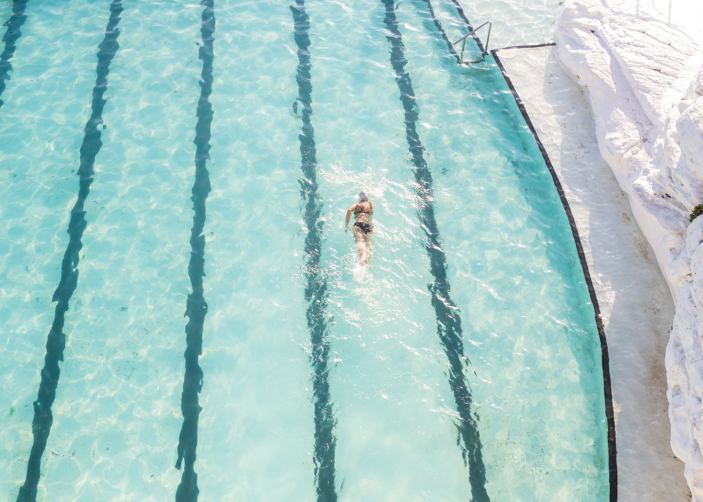 A person swims on their back in Bondi, a large, clear turquoise pool with distinct black lane markers from Francesca Owen. The pool is adjacent to a white, rocky area. The water glistens under sunlight, resembling the pure quality of fine art paper. A set of pool steps is visible in the top right corner.