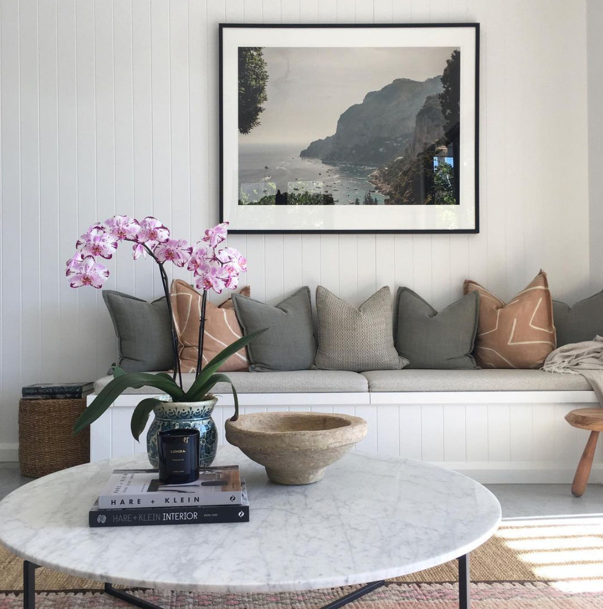 A cozy living room features a white bench adorned with assorted throw pillows. On the wall hangs "Capri Rooftops," a framed fine art print by Francesca Owen, and in front stands a white marble coffee table. The table displays an orchid plant, a candle, and books, adding a touch of elegance to the modern, inviting space.