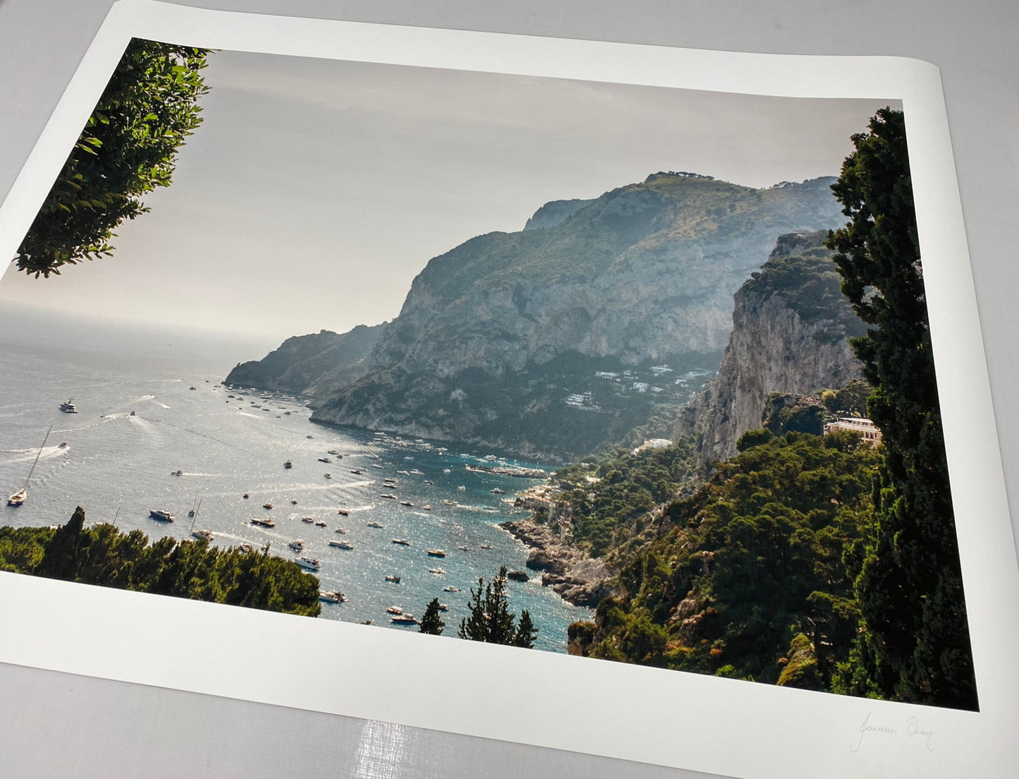 A scenic coastal landscape featuring rugged cliffs descending into the sea, dotted with boats. Surrounding the coastline are lush green trees and shrubbery, with the horizon fading into a hazy sky. This beautiful scene is available as fine art prints under the name "Capri Rooftops" by Francesca Owen, created with archival pigments and accompanied by a certificate of authenticity.