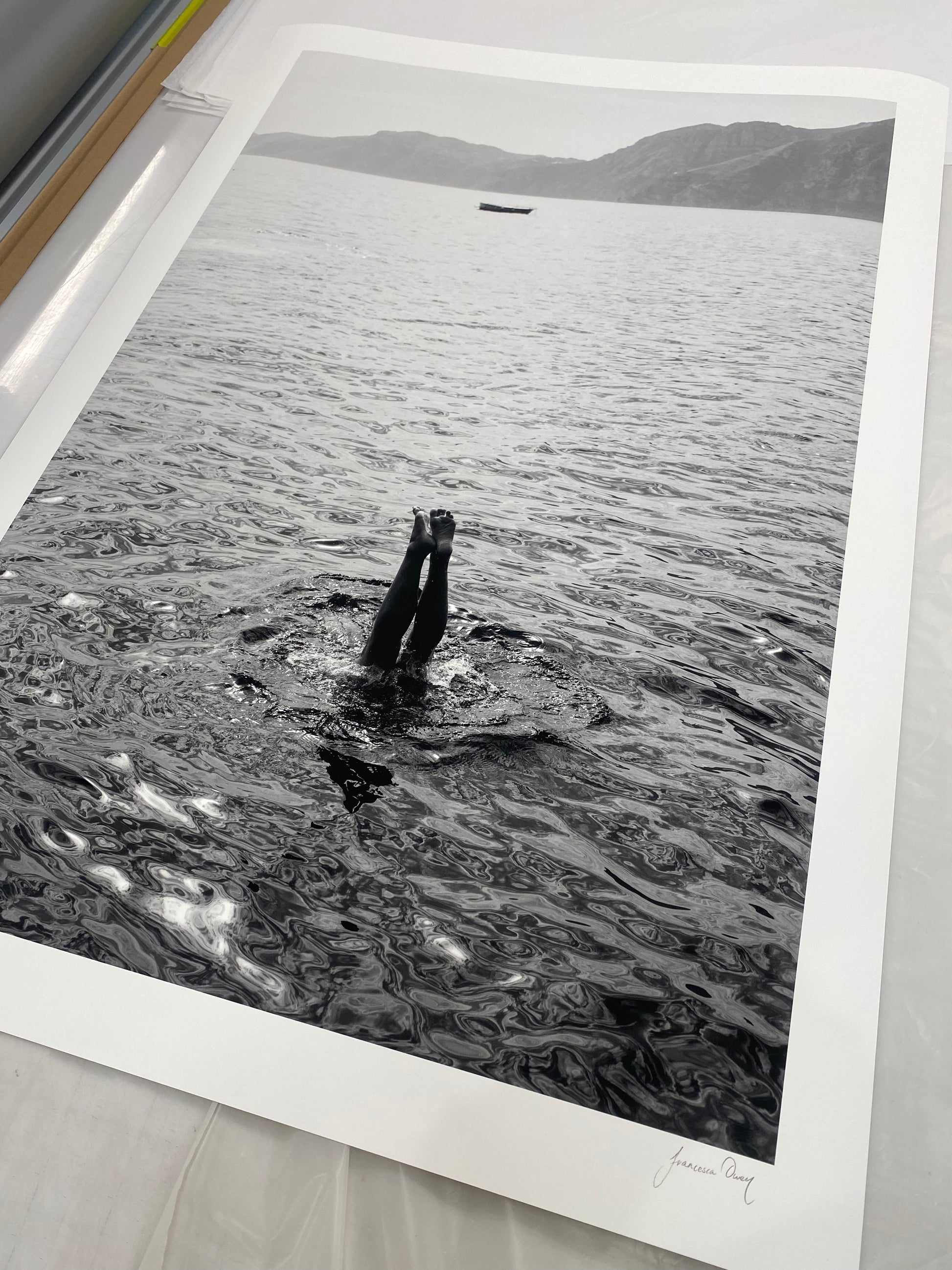 Capri Tippy Toes" by Francesca Owen captures a striking black and white image of a person performing a handstand in the water, with only their legs and feet visible above the surface. In the background, distant mountains and a solitary boat float on calm waters. This piece is available in custom sizes with archival pigments and includes a certificate of authenticity.