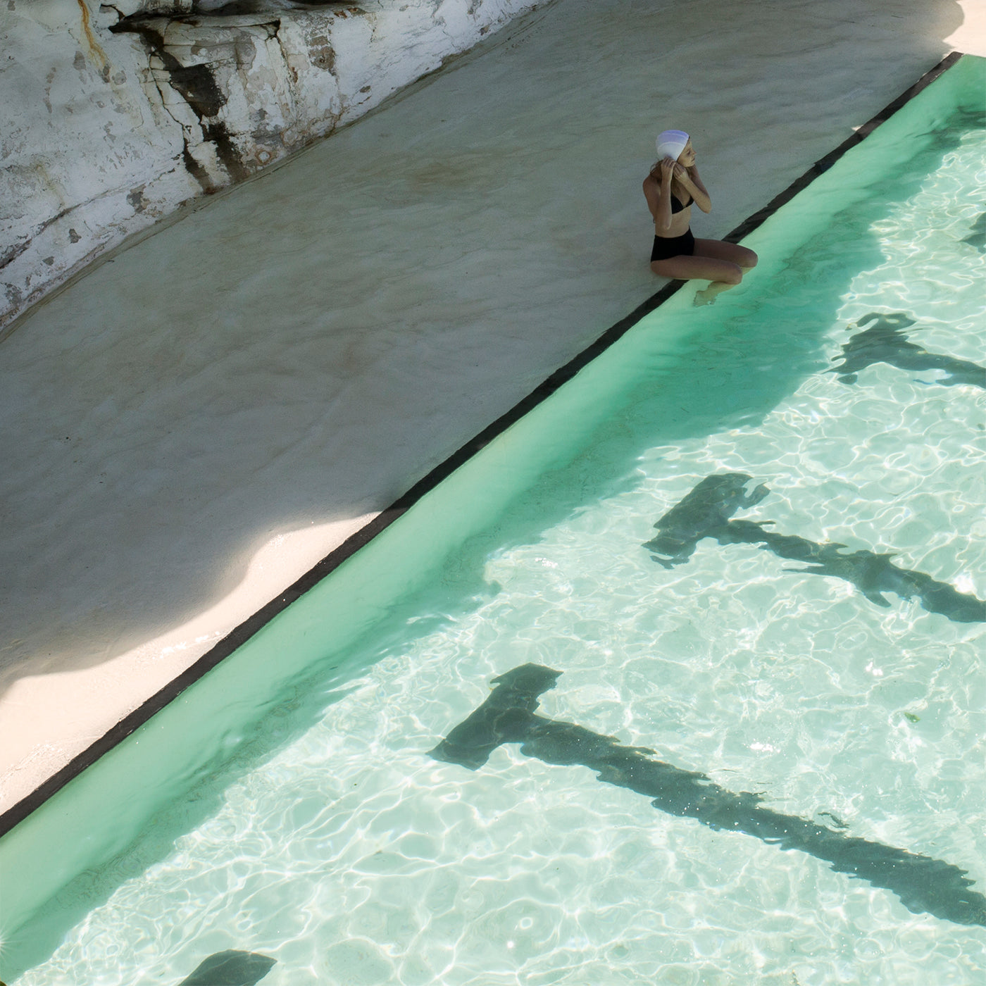 A person wearing a black swimsuit and white hat sits on the edge of a clear turquoise swimming pool, their feet dipping into the water. A rocky, sunlit wall serves as a striking backdrop. The scene evokes the timeless elegance of fine art prints, reminiscent of Francesca Owen's "Bondi Empty Lanes," with shadows cast by the pool's metal steps creating an almost archival pigments masterpiece.
