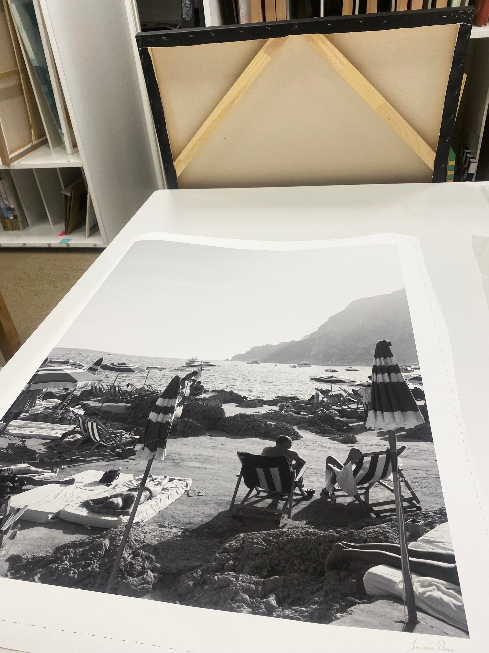 A black-and-white photograph titled "La Fontelina #2" by Francesca Owen depicts beachgoers lounging under umbrellas by the sea, with rocky cliffs in the background. The photo rests on a table in an art studio, printed on fine art paper using archival pigments, with bookshelves and a canvas visible in the background.