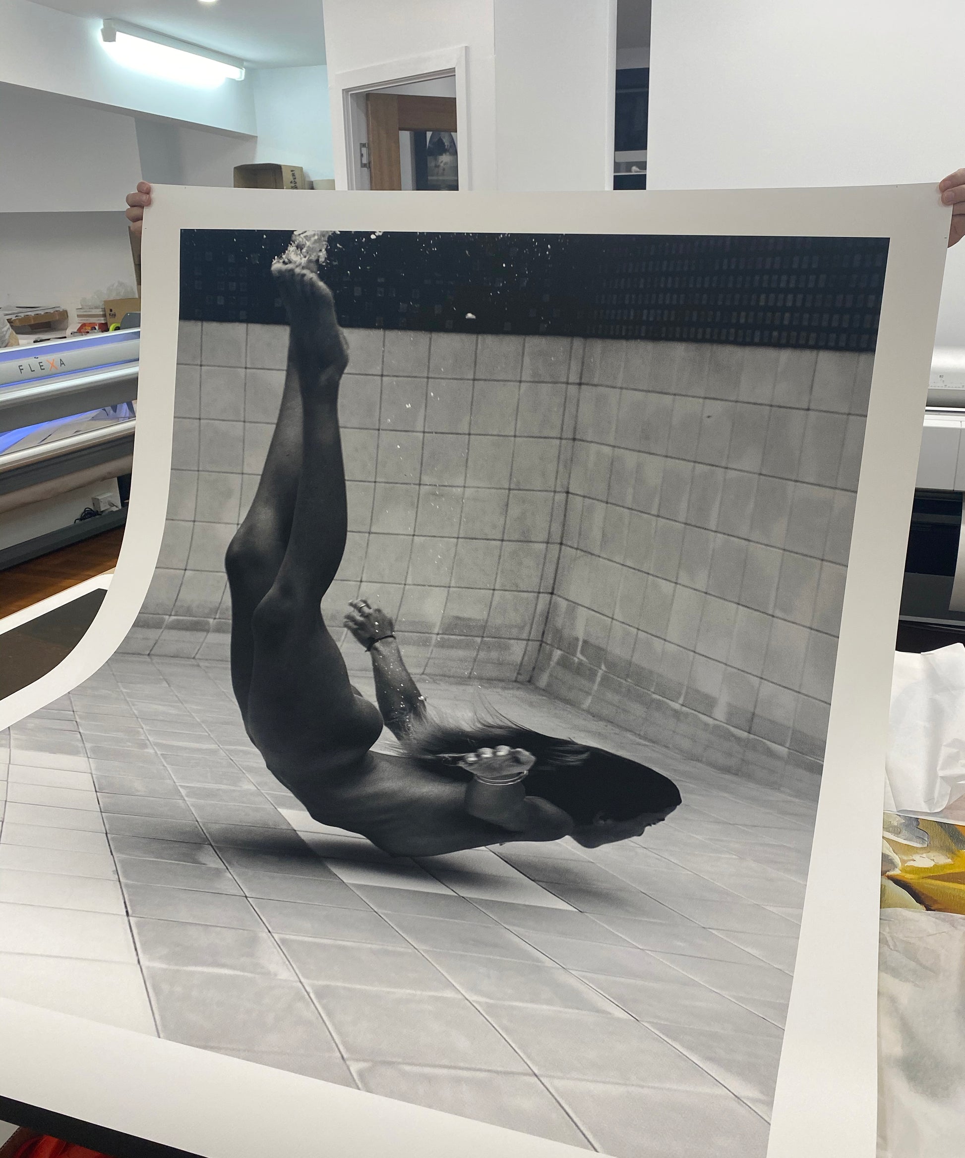 A large black-and-white photograph titled "Fall" by Francesca Owen is held up in a room. The image, printed on fine art paper, depicts a person underwater, upside down, with legs extended upward and arms outstretched. The tiled floor is visible, and bubbles surround the person, suggesting movement in the water.