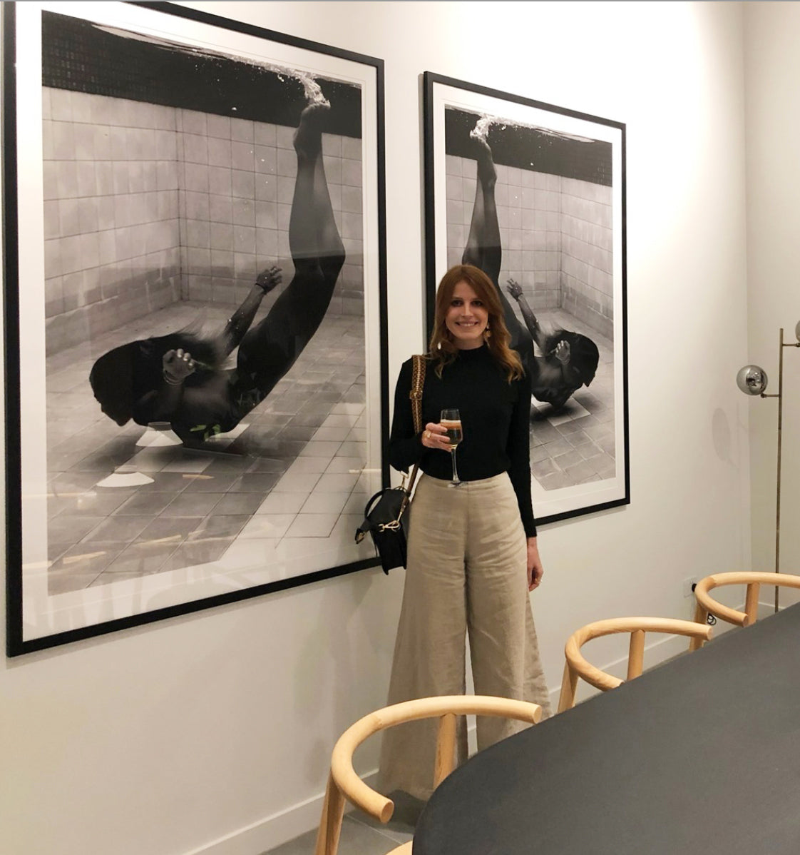 A woman in a Francesca Owen "Fall" black turtleneck and beige wide-legged pants stands smiling in a modern room. She holds a drink in one hand and a black purse in the other. Behind her, two large custom-framed photographs show a swimmer underwater in a pool. Wooden chairs are in the foreground.
