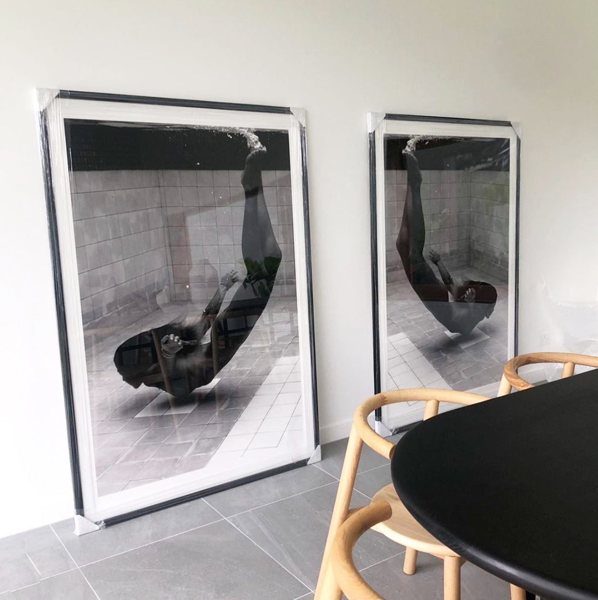 Two expertly crafted and custom-framed black-and-white art prints, from Francesca Owen's "Fall" collection, lean against a light gray wall. Each photograph depicts a person submerged underwater with legs extended upwards. In the foreground, there is a black table surrounded by light wood chairs.