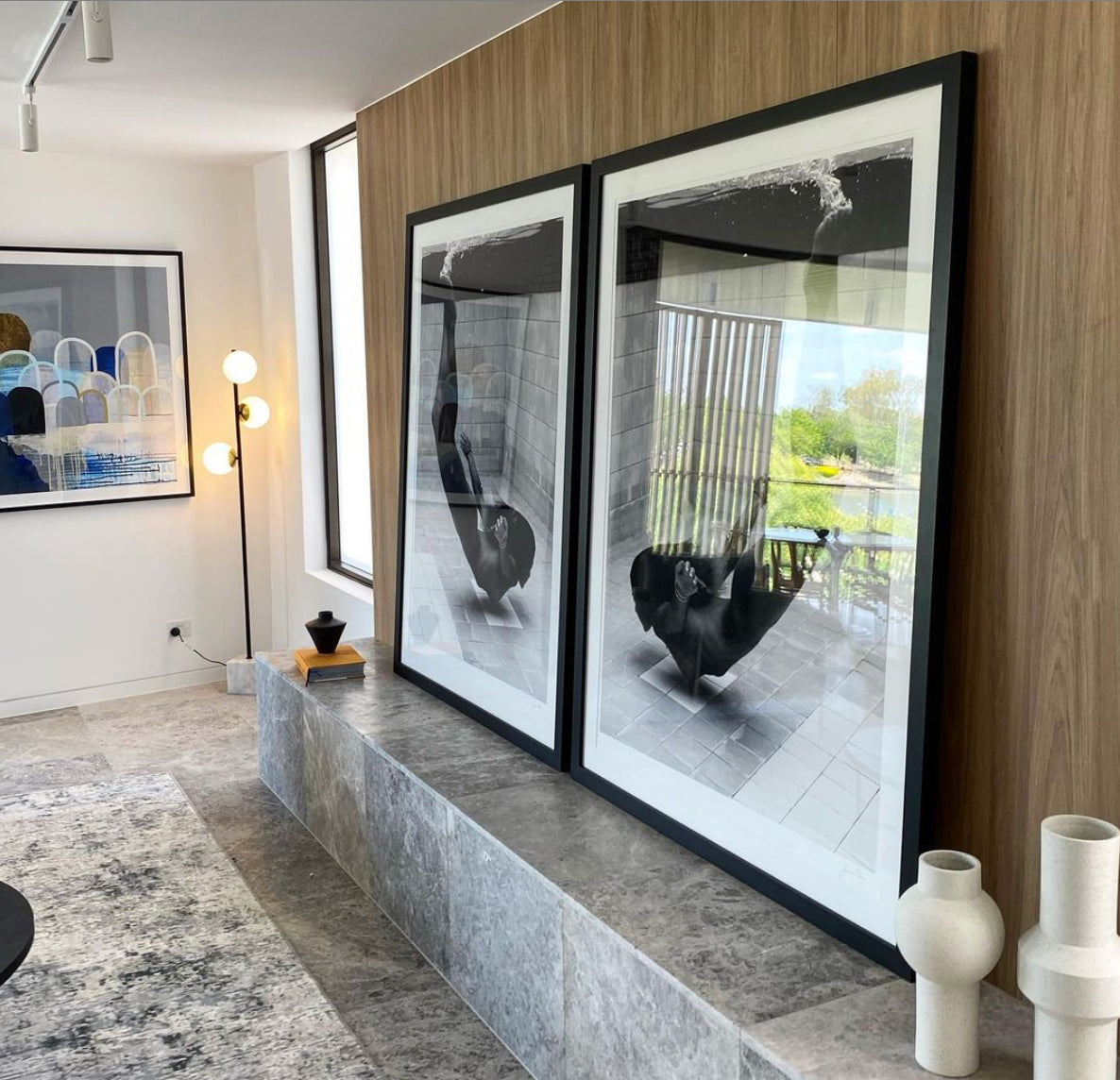The image showcases a contemporary living area featuring a stone-tiled bench set against a wooden-paneled wall. Two large, framed black-and-white art prints from the "Fall" series by Francesca Owen, depicting a person in a bathtub, rest against the wall. The room is accentuated with ceramic vases, a spherical lamp, and abstract artwork, all emphasizing the custom framing.