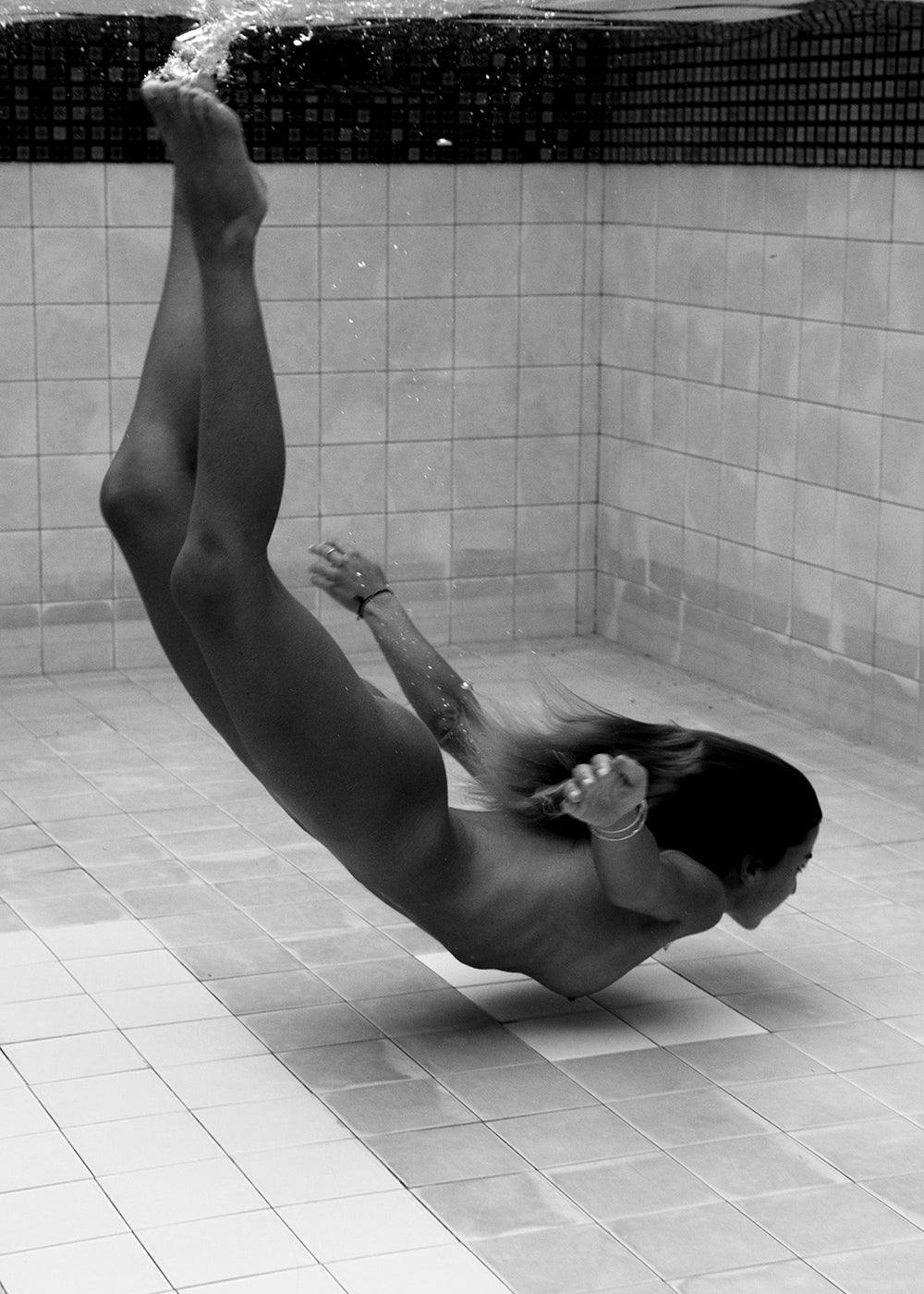 In the black-and-white photo "Fall" by Francesca Owen, a person in a swimsuit is captured underwater swimming gracefully in a pool, mid-dive, with arms extended forward and legs stretched back. Available as art prints on fine art paper with custom framing options, this artwork emphasizes the fluid motion and serene environment.